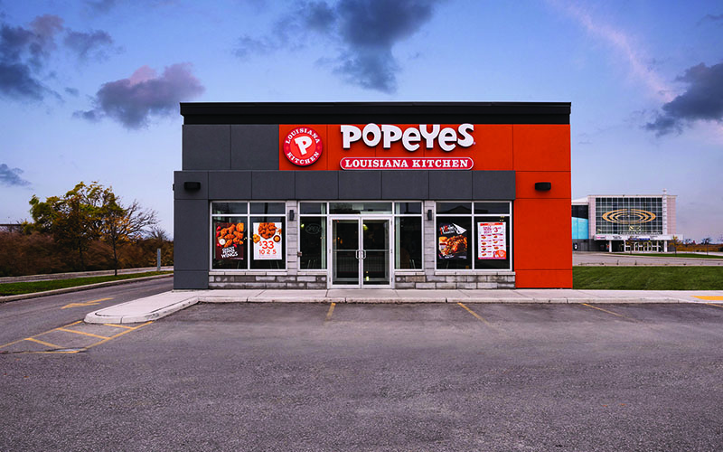 Exterior Rendering of a Popeyes Restaurant front of the building. Dark Grey brick with pops of Red along the top and down the right side of the building. Windows around the front door.